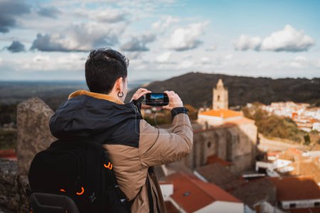 Foto de Joven fotografiando el panorama de una ciudad, viajando con su mochila, vista desde atrás, imagen en la pantalla del smartphone - Imagen libre de derechos
