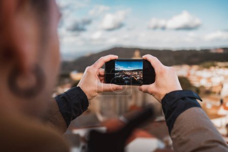 Téléchargez les photos : Jeune homme photographiant le panorama d'une ville, voyageant avec son sac à dos, vu de derrière, image sur l'écran du smartphone - en image libre de droit