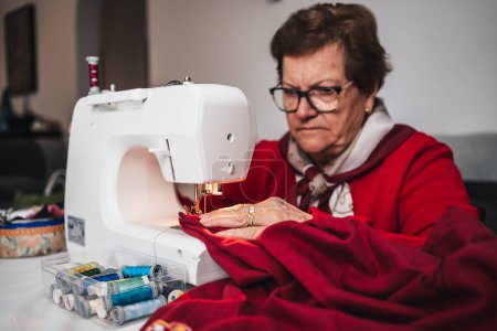 Téléchargez les photos : Senior femme en rouge et lunettes à la maison couture sur machine à coudre - en image libre de droit