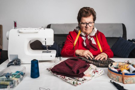 Foto de Costurera de moda en rojo con gafas de costura rodeada de máquina de coser, telas y paquetes de hilos para coser - Imagen libre de derechos