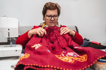 Téléchargez les photos : Femme plus âgée dans le salon de sa maison coudre un vêtement fait à la main faite par elle - en image libre de droit