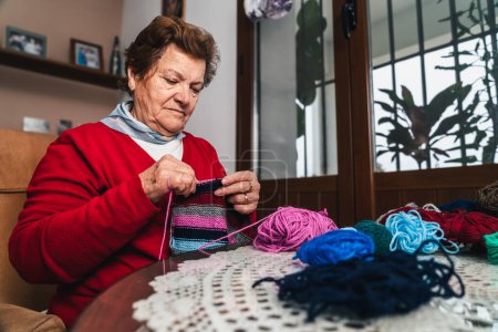 Foto de Mujer mayor tejiendo un trozo de lana para la ropa de manera artesanal, en la sala de estar de su casa junto a la ventana - Imagen libre de derechos