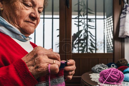 Photo for Close portrait of pretty and happy senior woman knitting a piece of woolen clothing with needles and colorful wool next to a window - Royalty Free Image