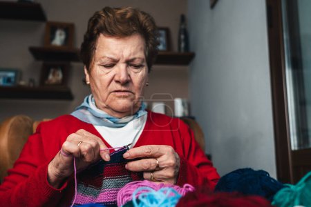 Téléchargez les photos : Portrait de jolie et heureuse femme âgée cousant un morceau de vêtements en laine avec des aiguilles et plusieurs boules de laine colorée dans le salon de sa maison - en image libre de droit