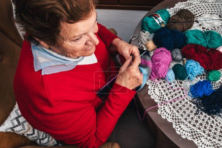Foto de Foto horizontal de la mujer mayor tejiendo con lana y agujas y mirando al lado sentado en la sala de estar en casa - Imagen libre de derechos