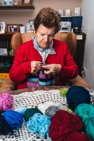 Foto de Retrato de mujer mayor y movimiento de sus manos cosiendo con agujas y lana de colores en un sofá en casa - Imagen libre de derechos