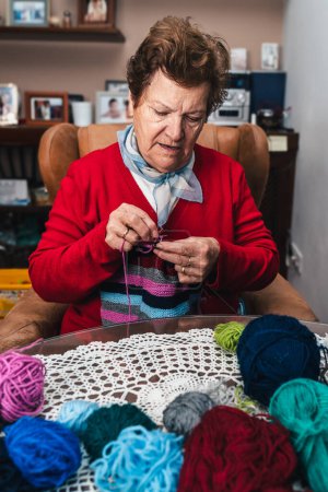 Photo for Portrait of senior woman and movement of her hands sewing with needles and colored wool on a sofa at home - Royalty Free Image