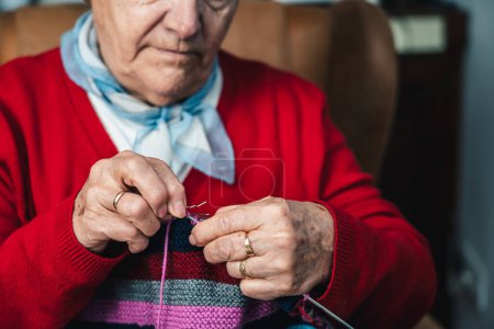 Téléchargez les photos : Portrait détail de la main de la femme âgée couture manœuvre tout en cousant un morceau de vêtements avec des aiguilles et de la laine colorée dans le salon de sa maison - en image libre de droit