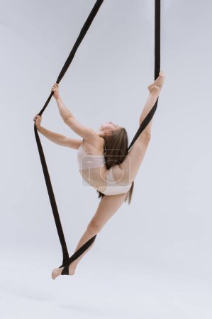 Chica gimnasta aérea demuestra estiramiento en cordel en trapecio acrobático. Atleta acrobático realiza colgando desde una altura.