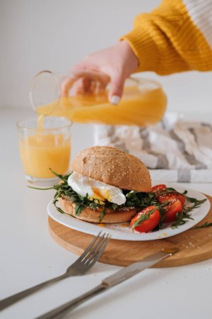 Foto de Sándwich de desayuno con huevo y rúcula y tomates cherry en la mesa, la mujer vierte jugo de naranja en un vaso - Imagen libre de derechos