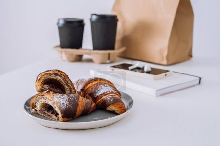 Foto de Croissants de chocolate con tazas de café y bloc de notas con smartphone y auriculares en mesa blanca, concepto de entrega de alimentos - Imagen libre de derechos