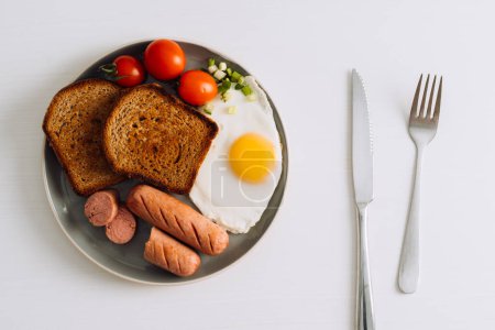 Foto de Placa de desayuno con cuchillo y tenedor en la mesa blanca, salchicha a la parrilla y tostadas de trigo integral con huevo frito y tomates cherry en el plato - Imagen libre de derechos