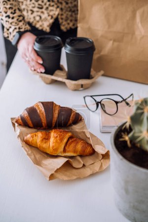 Foto de Deliciosos croissants marrones y chocolate y taza de café, mujer almorzando en el lugar de trabajo, concepto de entrega de alimentos - Imagen libre de derechos