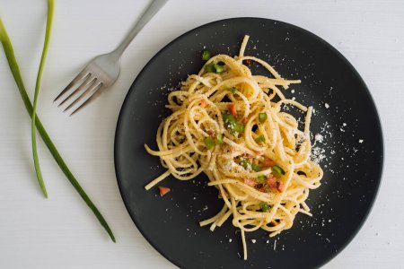 Foto de Pasta de espaguetis espolvoreada con queso parmesano y cebolla verde en un plato negro sobre una mesa blanca con tenedor - Imagen libre de derechos