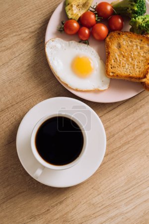 Foto de Desayuno casero con taza de café negro, huevo frito y tostadas con tomates y aguacate en el plato - Imagen libre de derechos