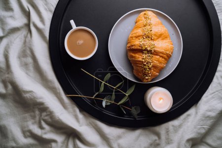 Photo for Flat lay of cup of cappuccino, croissant and scented candle with eucalyptus branch on tray on bed - Royalty Free Image