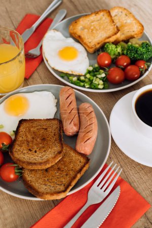 Foto de Desayuno inglés con taza de café negro y zumo de naranja, salchichas a la parrilla y tostadas de trigo integral con huevo frito y tomates cherry en el plato - Imagen libre de derechos