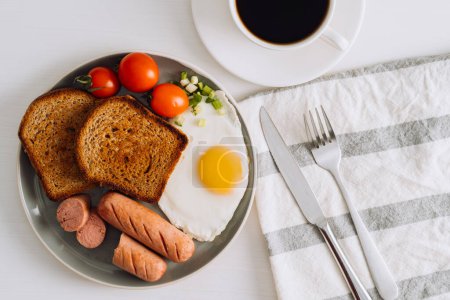 Foto de Plato de desayuno con taza de café negro, salchichas a la parrilla y tostadas de trigo integral con huevo frito y tomates cherry en el plato - Imagen libre de derechos