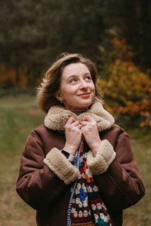 Foto de Mujer caucásica feliz en chaqueta marrón está de pie en el bosque de otoño y alegremente mirando hacia arriba - Imagen libre de derechos