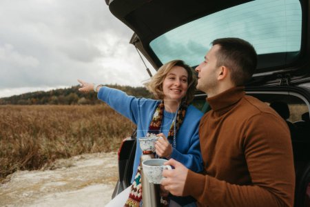 Foto de Feliz joven mujer y hombre sentado en el maletero abierto del coche mientras viaja en otoño, concepto de viaje por carretera - Imagen libre de derechos