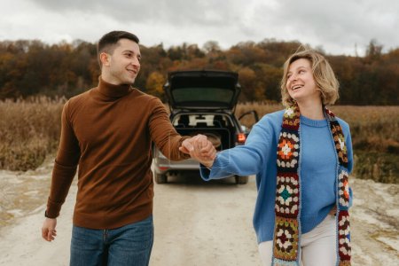 Téléchargez les photos : Couple souriant tenant la main, se promenant sur la route de campagne avec voiture en arrière-plan, homme et femme heureux en voyage - en image libre de droit