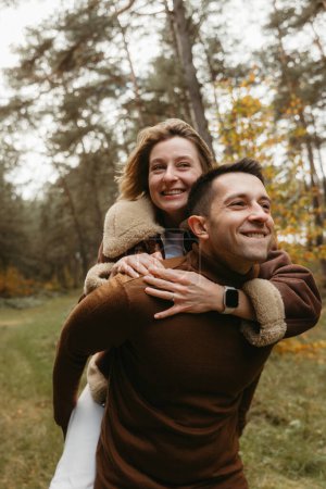 Foto de Pareja adulta divirtiéndose en el parque de otoño al aire libre, hombre feliz está llevando a la mujer en su espalda en el bosque - Imagen libre de derechos