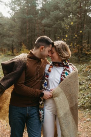 Foto de Un hombre y una mujer cubiertos de una manta acurrucados el uno al otro en medio del bosque, una pareja disfrutando de tiempo juntos fuera de la ciudad - Imagen libre de derechos