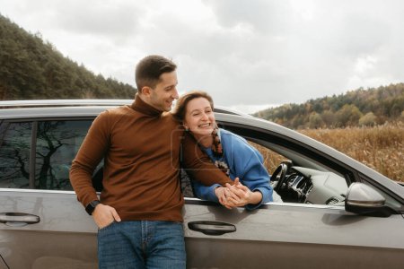 Foto de Hombre feliz inclinado al coche y cogido de la mano de la mujer alegre que se sienta dentro, pareja adulta disfrutando de viaje por carretera en otoño - Imagen libre de derechos