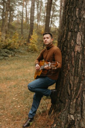 Foto de Hombre adulto caucásico toca la guitarra apoyada en el árbol en el parque de otoño - Imagen libre de derechos