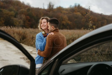 Foto de Abrazando al hombre y una mujer de pie junto a un coche en el camino de tierra y sonriendo, pareja disfrutando de viaje en carretera - Imagen libre de derechos