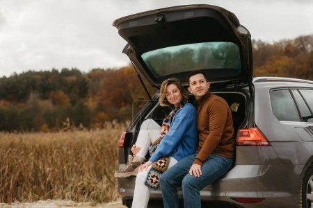 Foto de Feliz joven mujer y hombre sentado en el maletero abierto del coche mientras viaja en otoño y mirando hacia otro lado, concepto de viaje por carretera - Imagen libre de derechos