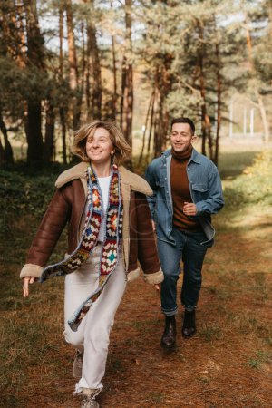Foto de Pareja feliz se divierte en medio del bosque, mujer sonriente está corriendo con su marido en un parque de otoño - Imagen libre de derechos