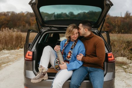 Foto de Feliz pareja adulta sentada en el maletero abierto del coche mientras viaja en otoño, concepto de viaje por carretera, hombre abrazando a la mujer - Imagen libre de derechos