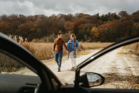 Foto de Hombre feliz y una mujer están caminando por un camino rural junto a su coche cogidos de la mano, pareja adulta en viaje por carretera en otoño - Imagen libre de derechos