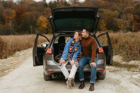 Foto de Feliz joven mujer y hombre sentado en el maletero abierto de su coche mientras viaja en otoño y mirando hacia otro lado, concepto de viaje por carretera pareja - Imagen libre de derechos
