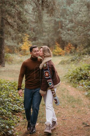 Foto de Una mujer está besando a un hombre en la mejilla mientras camina por el bosque de otoño, pareja feliz pasando tiempo juntos - Imagen libre de derechos