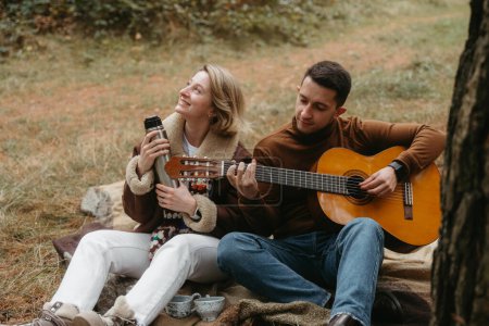 Foto de Pareja adulta sentada en una manta en el parque de otoño, un hombre tocando la guitarra para una mujer feliz - Imagen libre de derechos
