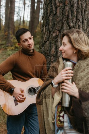 Foto de Un hombre está tocando una guitarra para mujer en el bosque en otoño, pareja adulta pasando tiempo juntos - Imagen libre de derechos
