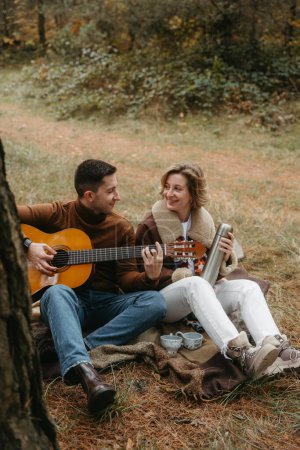 Foto de El hombre está tocando la guitarra para la mujer feliz, pareja caucásica adulta sentada en una manta en el parque de otoño - Imagen libre de derechos