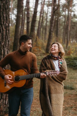 Téléchargez les photos : Un homme en jean joue de la guitare pour une femme couverte de plaid. Ils sont entourés d'arbres et d'un paysage naturel, tous deux souriants - en image libre de droit