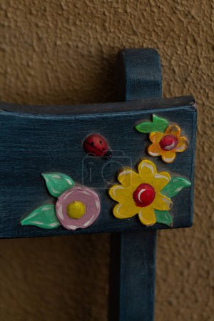 Close-up of a part of a blue wooden bench with old childish decorations of colorful flowers and a ladybug with a brown wall background