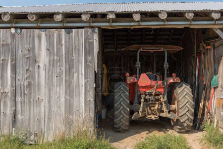 Tractor parked in a shed with farm tools and equipment on a sunny late afternoon. High quality photo