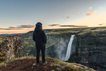 Téléchargez les photos : Rear view of man in winter coat standing and looking at Majestic Haifoss waterfall flowing in volcanic canyon among the Icelandic Highlands on summer at Iceland - en image libre de droit