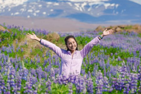 Foto de Atractiva mujer asiática feliz de pie y sonriendo en el campo de flores de altramuz en verano en Islandia - Imagen libre de derechos