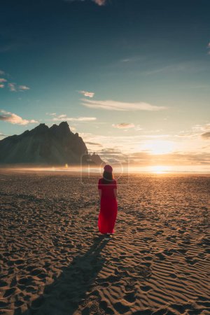 Photo for Beautiful view of a woman in red dress standing with sunlight glowing in Vestrahorn mountain during sunrise on black sand beach in Stokksnes peninsula at Iceland - Royalty Free Image