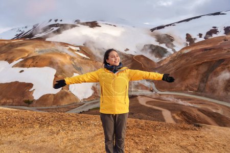 Foto de Joven mujer asiática alegre en chaqueta amarilla de pie en la cordillera Kerlingarfjoll en la zona geotérmica en verano en las tierras altas de Islandia - Imagen libre de derechos