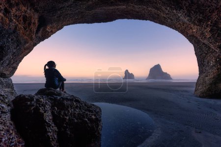 Female tourist sitting on rock in stone cave with Archway islands on Wharariki beach in the morning at Cape Farewell, New Zealand