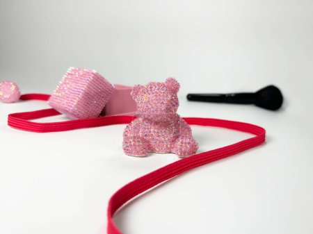 Ein funkelnder rosa Teddybär mit glänzenden Strasssteinen, der auf weißem Hintergrund ruht. Ein roter Streifen umgibt diese verspielten Objekte und schafft einen ästhetischen Reiz.