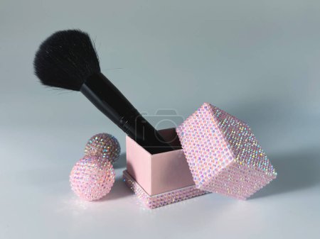 La imagen cuenta con un cepillo de maquillaje con un mango negro y cerdas oscuras descansando en una caja rosa abierta adornada con adornos brillantes.