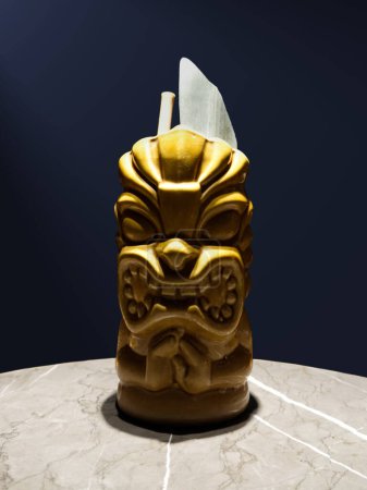 Goldener Tiki-Becher mit bedrohlichem Gesicht sitzt auf einer Marmoroberfläche, dessen glänzende Außenseite vor dem dunklen Hintergrund kontrastiert. Der Becher enthält einen Cocktail, garniert mit einem grünen Blatt und einem Strohhalm. 